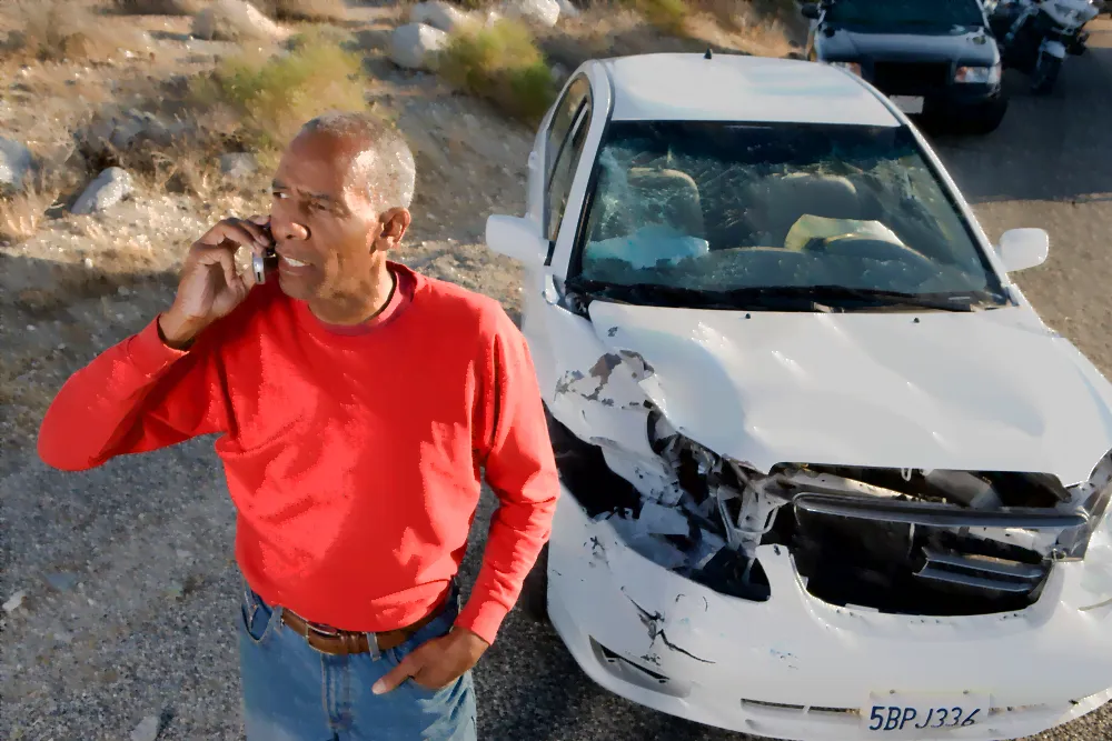 Man on his cell phone while his car is behind him damaged after a car accident.