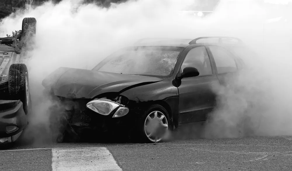 Black car with steam all around it. the car is damaged in the front and next to a flipped over car.