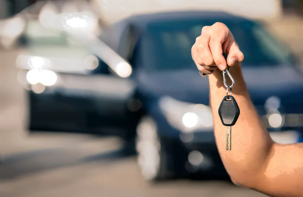 Keys being held up by a hand and a car in the back round.