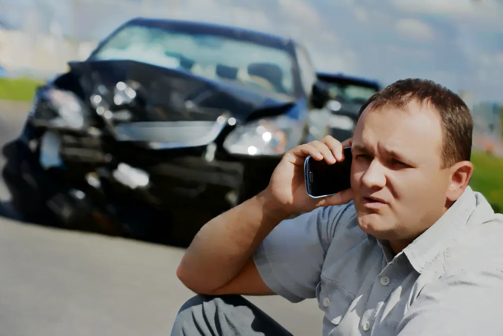 man on his cell phone after a car accident. he is on the ground calling and his damaged car his behind him.