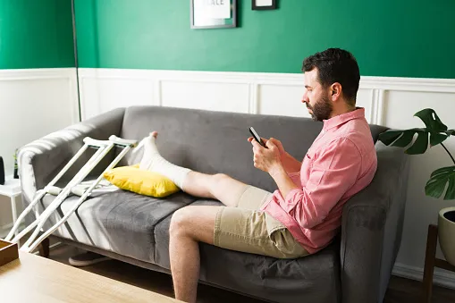 Injured man sitting on the couch with his leg in a cast, crutches by his side, and looking for a personal injury lawyer on his phone.