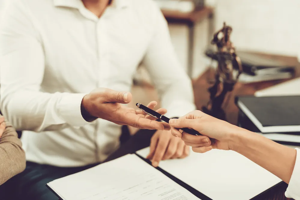 a person sitting at a desk being handed a pen and about to sign some papers in front of them.